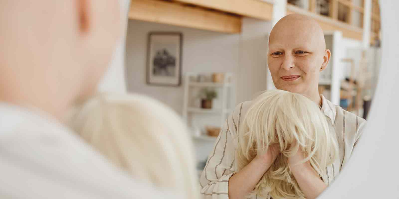 Frau ohne Haare steht mit einer blonden Perücke vor einem Spiegel und betrachtet diese